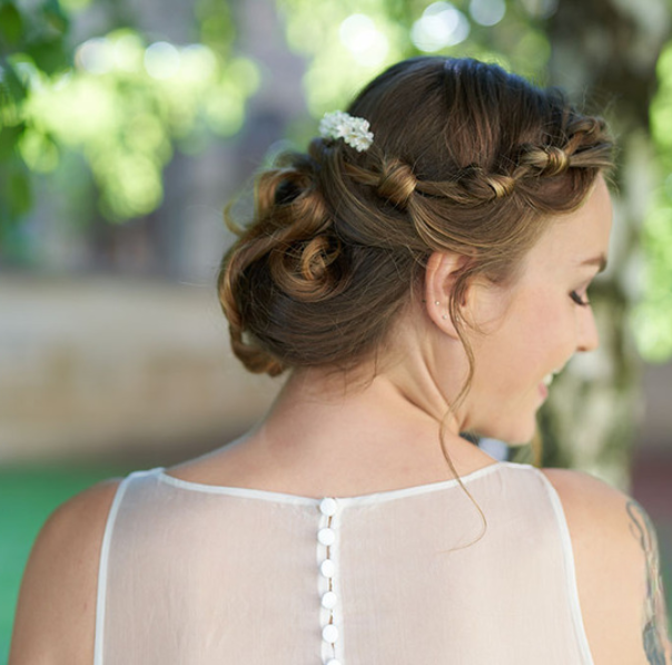 Núvia amb recollit baix trenat i detall floral al cabell, amb vestit blanc d’esquena amb botons, en un entorn natural.