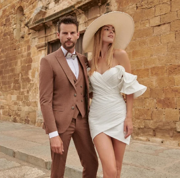 Parella vestida per a un casament a Girona: el nuvi amb vestit formal d’ONIX i la núvia amb vestit blanc curt i barret, sobre un fons de pedra.
